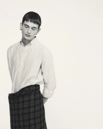 Model with short dark hair wearing a striped button-down shirt and plaid skirt against a plain light background