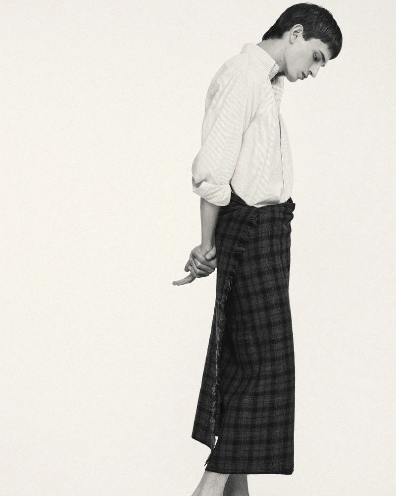 Profile view of a male model in a white shirt and dark plaid kilt against a plain background for Frere.