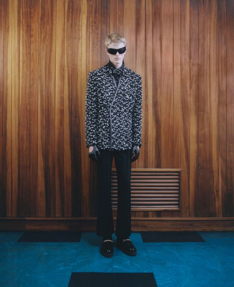 Fashionable adult in patterned jacket, black pants, and sunglasses poses against a wood panel wall in a studio setting.