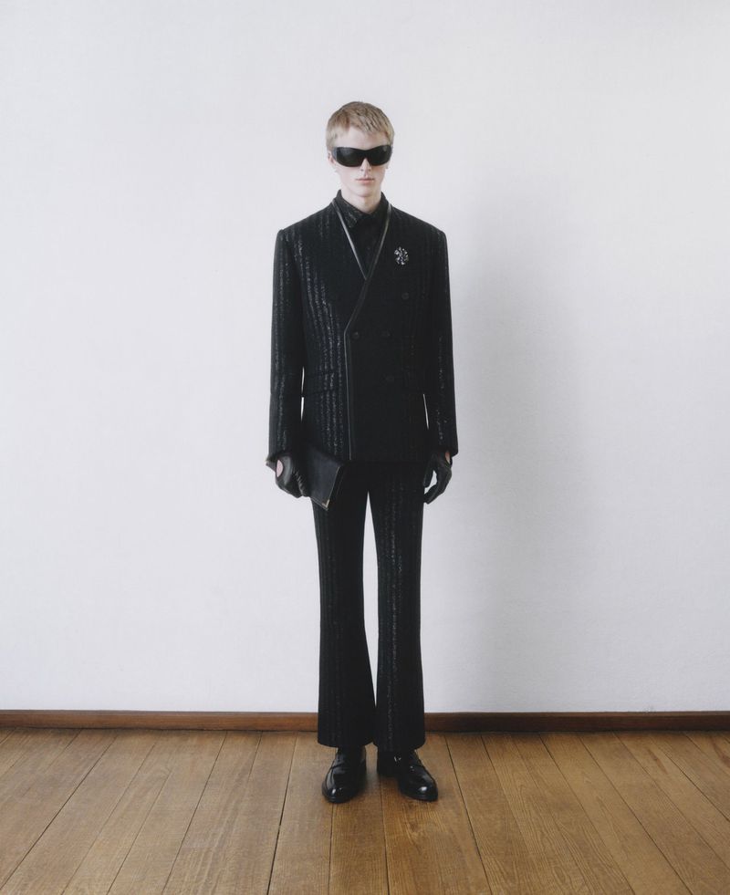 A man poses in a black double-breasted suit with sunglasses and a leather clutch against a stark white wall with a wood floor.