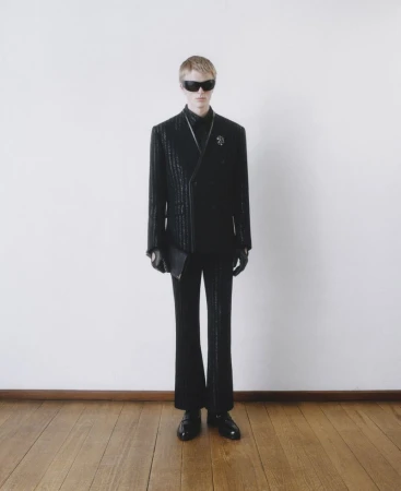 A man poses in a black double-breasted suit with sunglasses and a leather clutch against a stark white wall with a wood floor.
