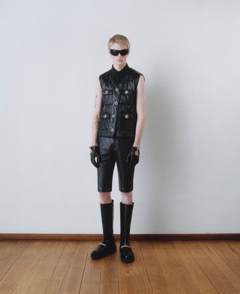 A fair-skinned male model poses in a black leather vest, shorts, gloves, sunglasses, and boots against a white wall on a wood floor.