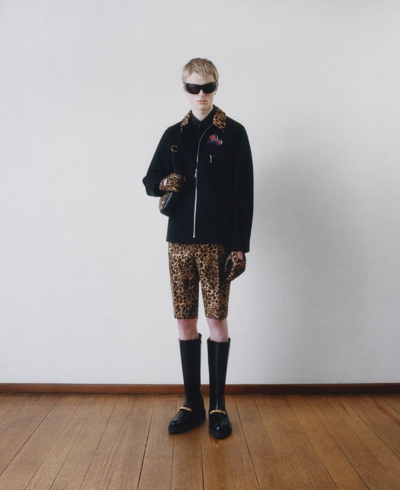 Full-length studio shot of a young adult wearing a black jacket, leopard-print shorts, sunglasses, and black boots against a white wall.