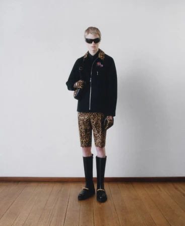 Full-length studio shot of a young adult wearing a black jacket, leopard-print shorts, sunglasses, and black boots against a white wall.