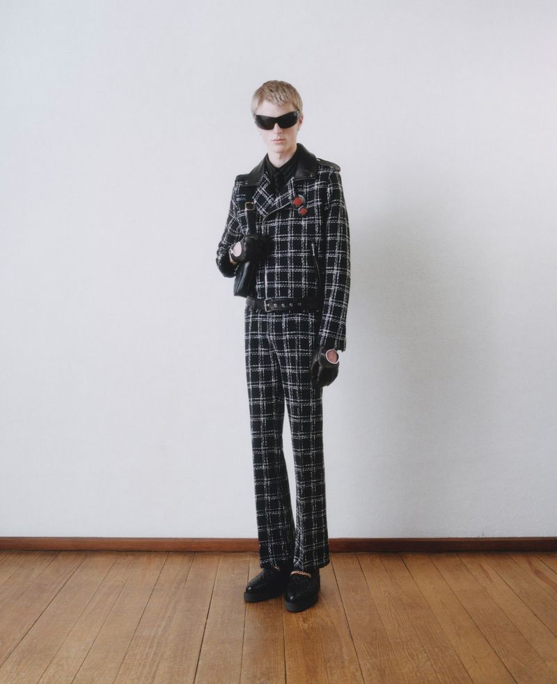 Fashionable young adult posing in a black and white checkered suit with dark sunglasses in a studio setting. Full-length shot, clean background.