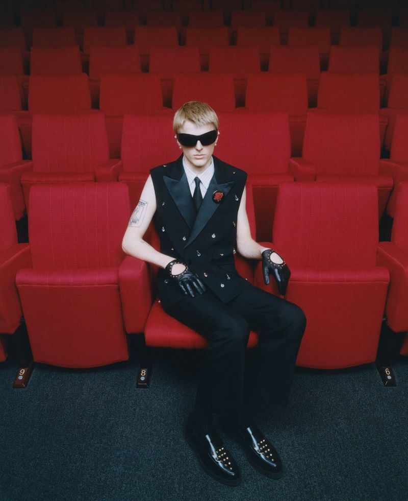 Man in black vest and sunglasses sits in red theatre seat, looking cool and stylish in an entertainment venue. Red chairs background.