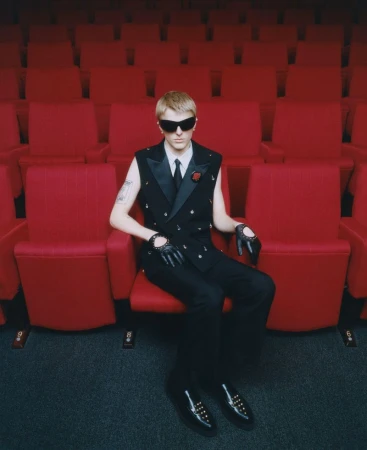 Man in black vest and sunglasses sits in red theatre seat, looking cool and stylish in an entertainment venue. Red chairs background.