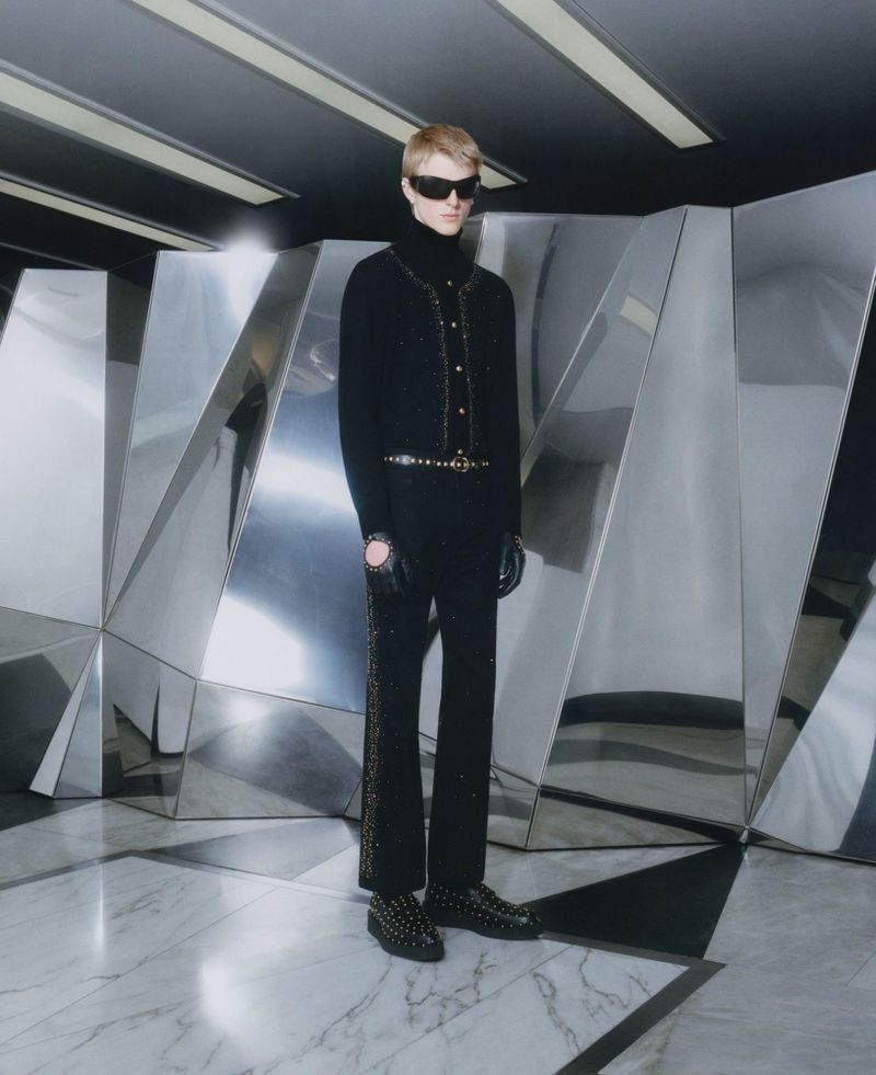 Fashion shot of a fair-skinned adult male wearing a black outfit, sunglasses, and studded boots, posing against a mirrored geometric wall.