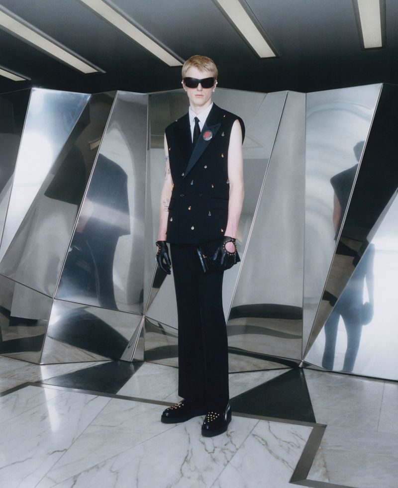 A person wearing sunglasses, a sleeveless blazer, and black pants stands in a geometric hall with metallic paneling and marble floors.