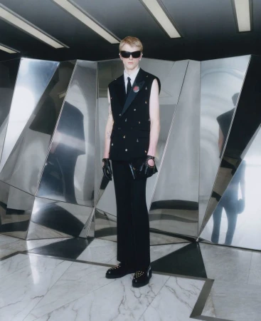A person wearing sunglasses, a sleeveless blazer, and black pants stands in a geometric hall with metallic paneling and marble floors.