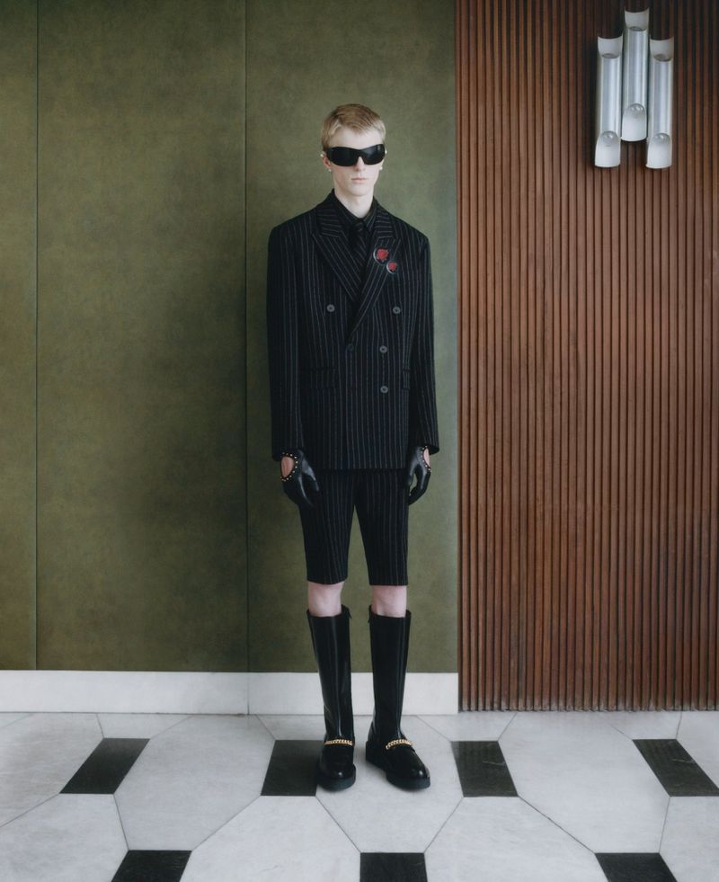 A fair-skinned young adult stands wearing a pinstripe suit with short pants, knee-high boots, sunglasses and gloves, posing in an indoor setting.