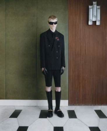 A fair-skinned young adult stands wearing a pinstripe suit with short pants, knee-high boots, sunglasses and gloves, posing in an indoor setting.