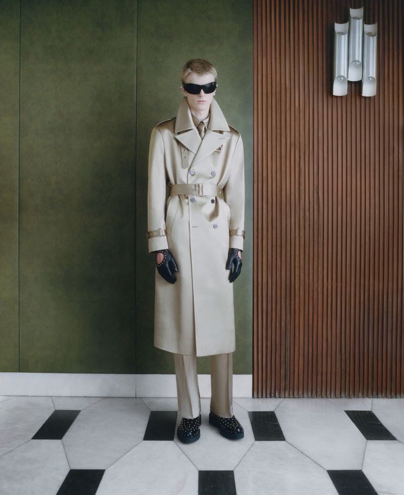 A blonde man wearing a tan trench coat, black studded loafers, and sunglasses stands in front of a paneled wall with tile flooring.
