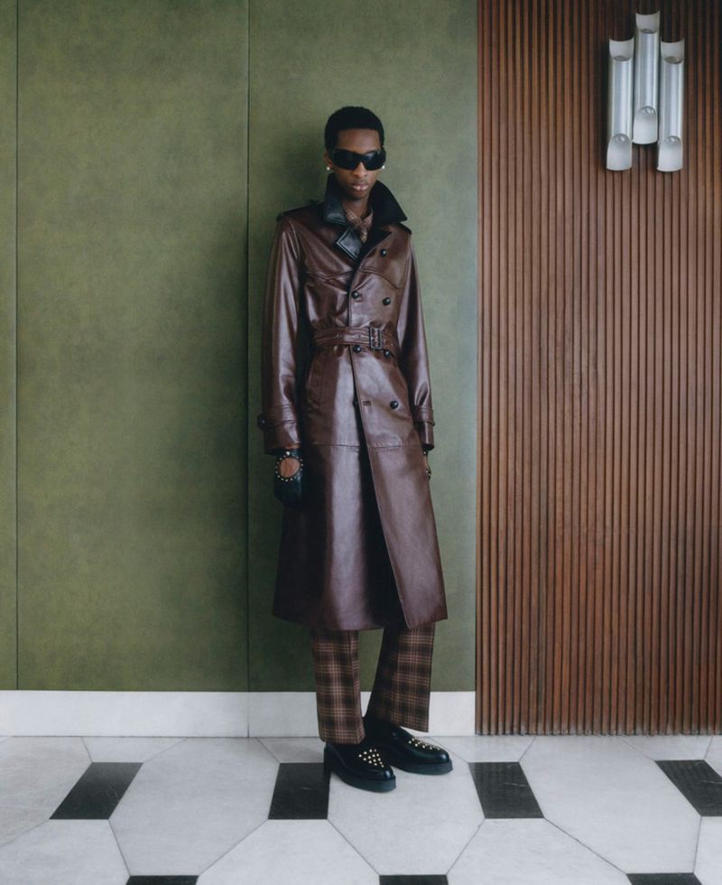 Man in a brown leather trench coat stands against a green and brown panel wall, showcasing fashion with style and sophistication.