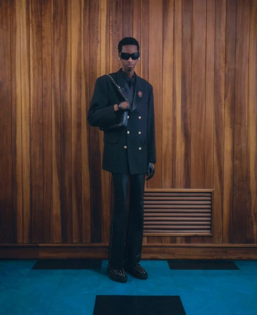 Full-length portrait of an adult male wearing a black double-breasted suit with sunglasses in front of a brown wood panel wall.