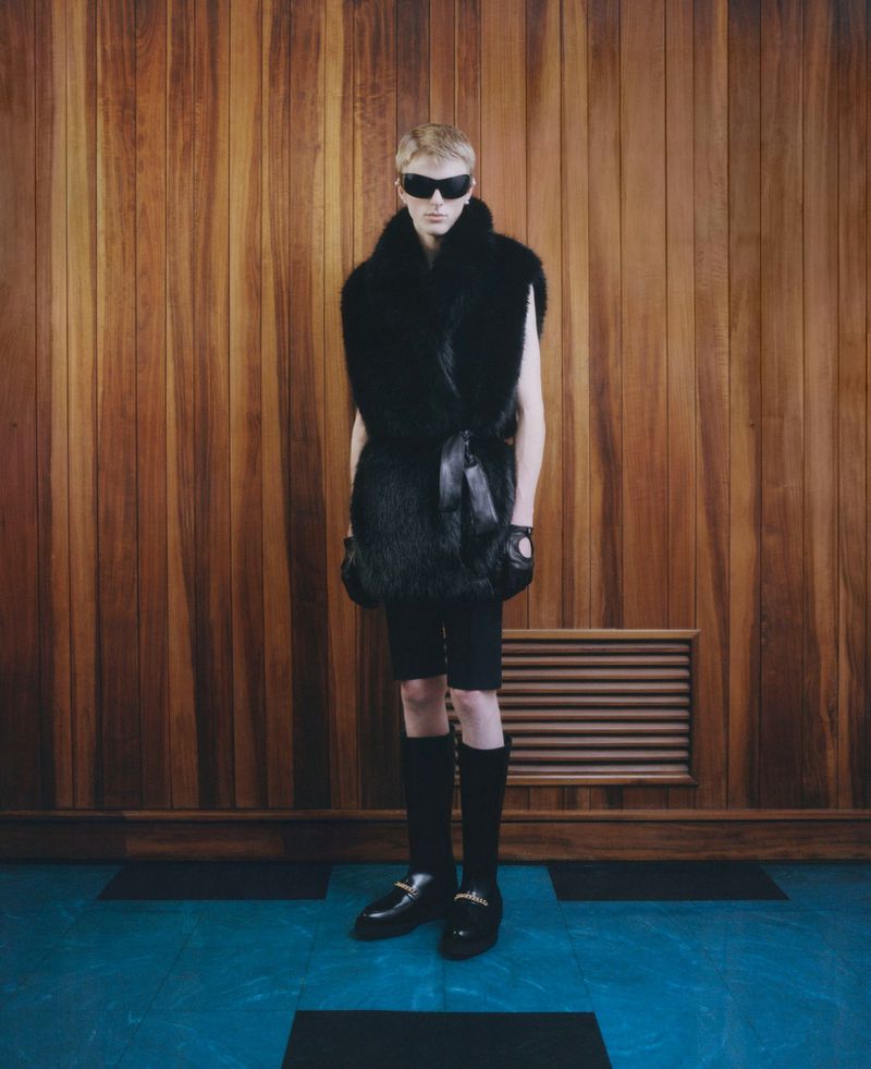 A fair-skinned person in dark sunglasses, a fur vest, and knee-high socks stands in a wooden panelled room with a painted blue floor.