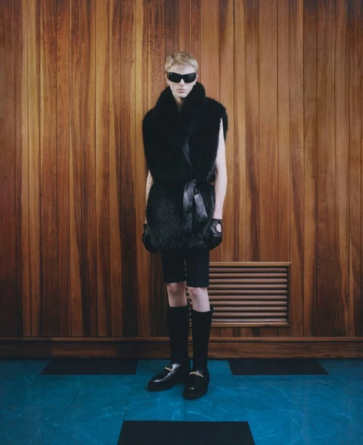 A fair-skinned person in dark sunglasses, a fur vest, and knee-high socks stands in a wooden panelled room with a painted blue floor.