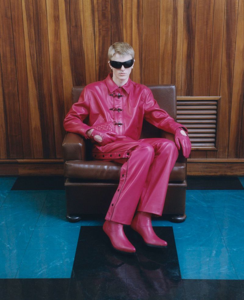 A fair-skinned adult wearing a bright-pink leather outfit and black sunglasses sits in a leather chair against a wood-panelled wall.