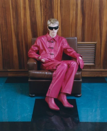 A fair-skinned adult wearing a bright-pink leather outfit and black sunglasses sits in a leather chair against a wood-panelled wall.