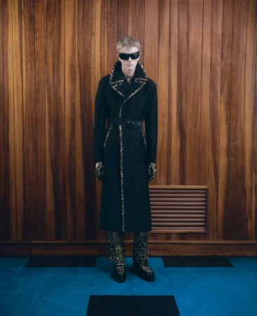 Full-length studio shot of an adult male fashion model wearing a black trench coat, leopard print pants, and sunglasses against a wood wall.
