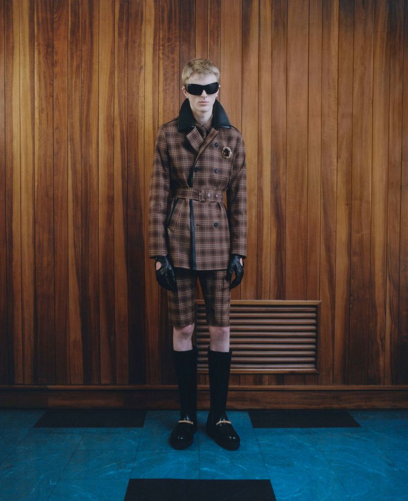 A fair-skinned adult man in a brown plaid suit, black gloves, and dark sunglasses stands in front of a wood-paneled wall.