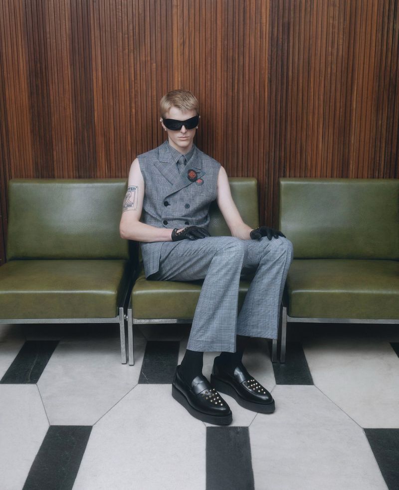 A blond male model poses on a green bench indoors, wearing a gray plaid suit vest, pants, black socks, loafers, and sunglasses.