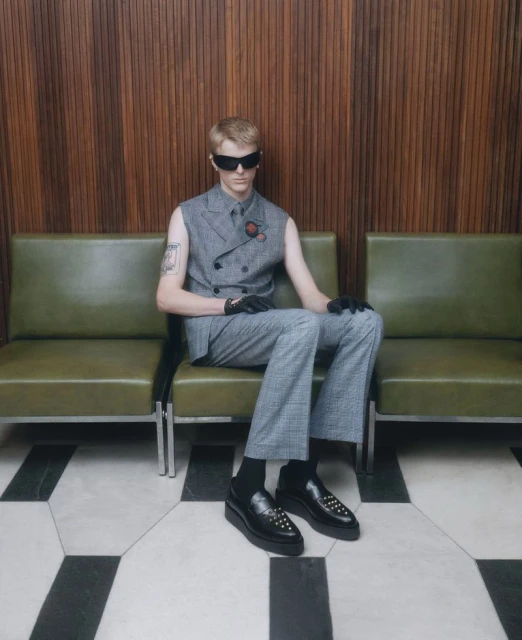 A blond male model poses on a green bench indoors, wearing a gray plaid suit vest, pants, black socks, loafers, and sunglasses.