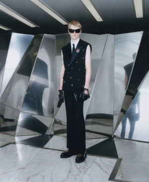 Model wearing a black sleeveless double-breasted suit vest, sunglasses, cut-out gloves, and studded shoes in front of a mirrored wall.