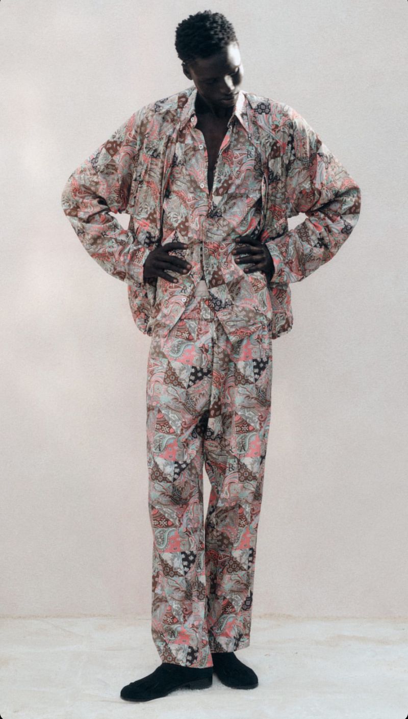 Adult Black man stands with hands on hips in matching pink patterned, long-sleeved shirt and pants against a light neutral background.