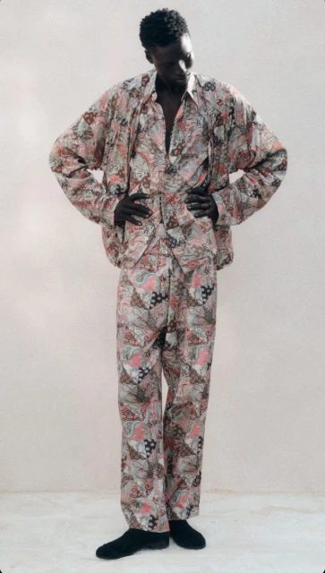 Adult Black man stands with hands on hips in matching pink patterned, long-sleeved shirt and pants against a light neutral background.