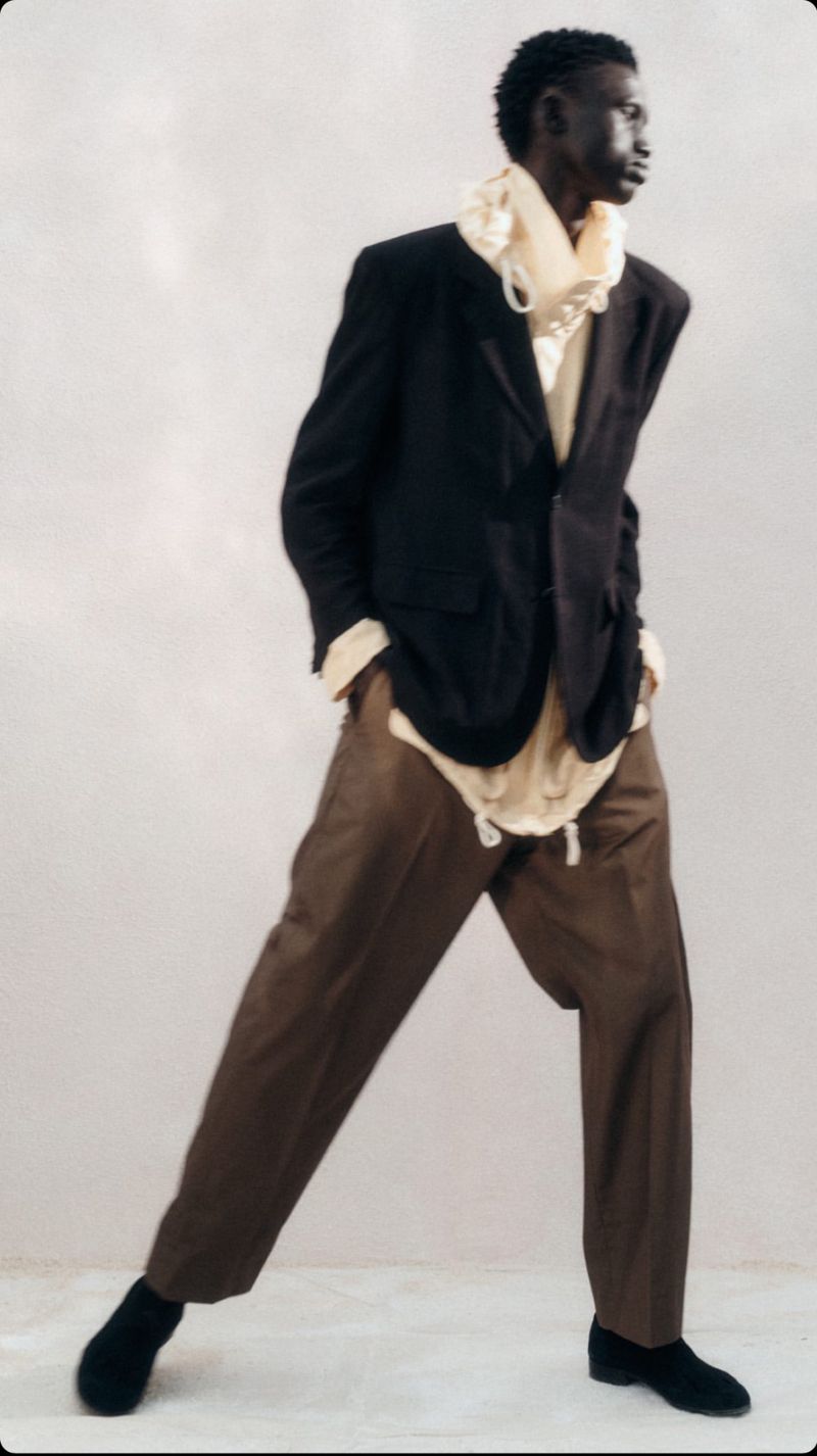 A Black male model strikes a dynamic pose in a black blazer, cream top, brown pants, and black shoes, set against a neutral background.