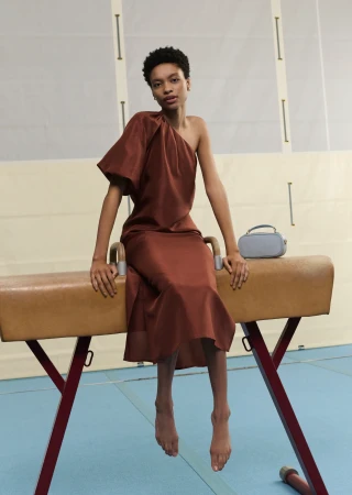 A woman wearing a copper-colored, one-shoulder dress sits on a gymnastics horse in an indoor gymnasium setting with soft lighting.