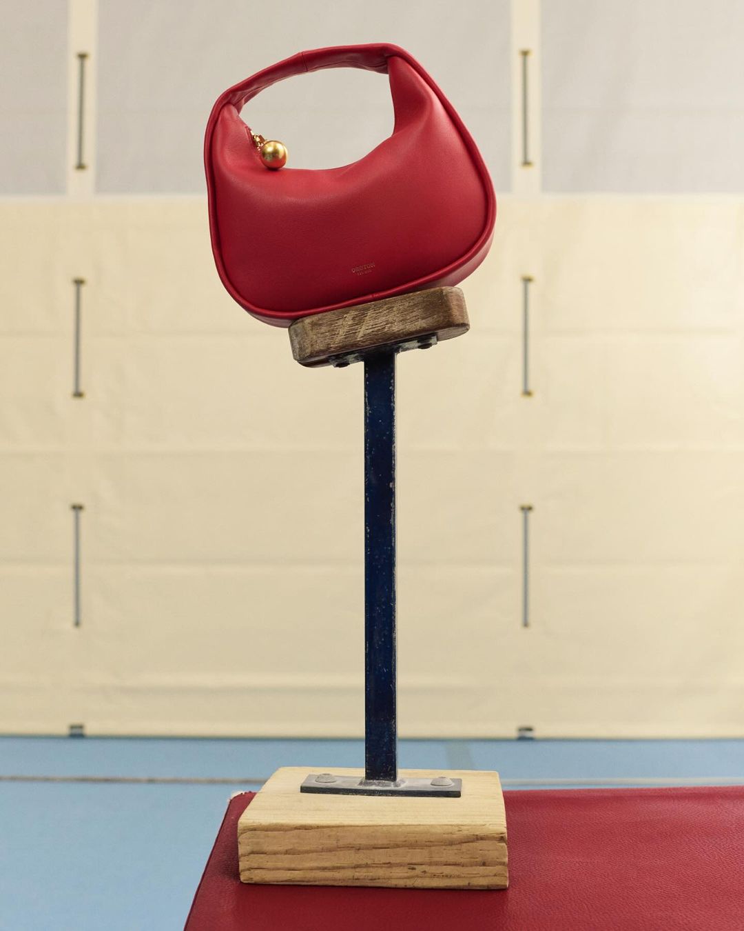 Still life of a red leather handbag with golden metal hardware perched on a blue and wooden stand in a minimalist photo studio.