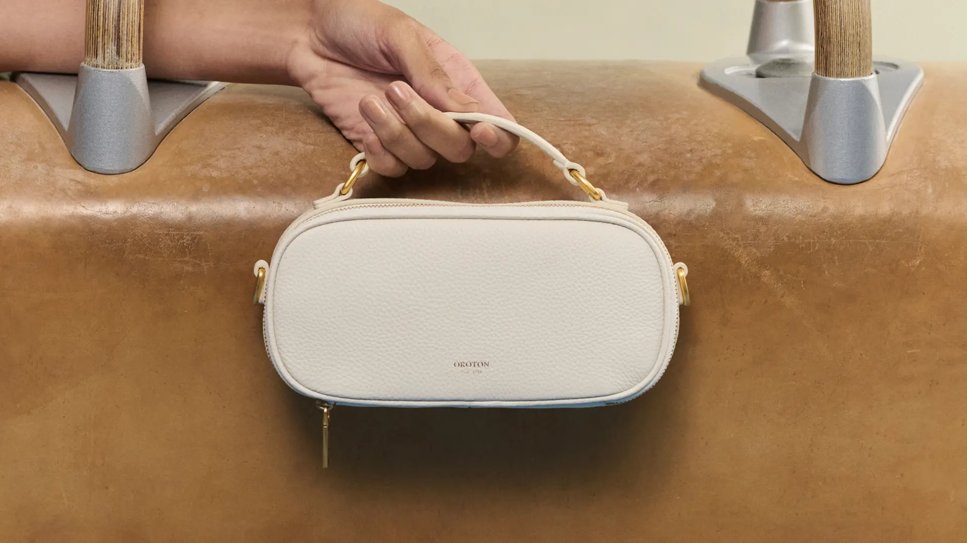 Close-up of a hand holding a white Oroton leather handbag with gold hardware, against a light brown leather pommel horse.