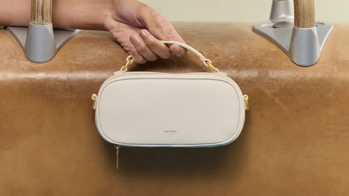 Close-up of a hand holding a white Oroton leather handbag with gold hardware, against a light brown leather pommel horse.