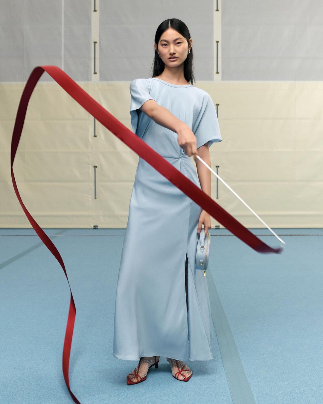 An Asian woman in a pale blue dress poses indoors with a red ribbon. She holds a small, boxy handbag in her left hand.