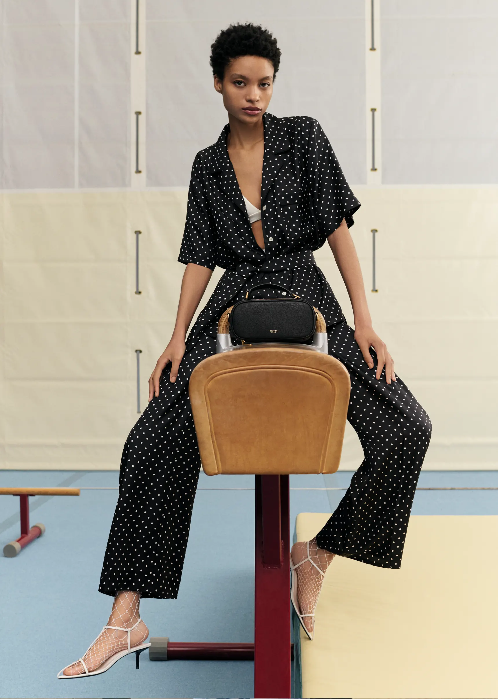 Elegant woman in polka dot top and pants sits on gym equipment with black purse and white heels, studio shot, fashion editorial.