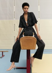 Model in a black polka dot jumpsuit sitting on a gymnasium vaulting horse with a handbag.