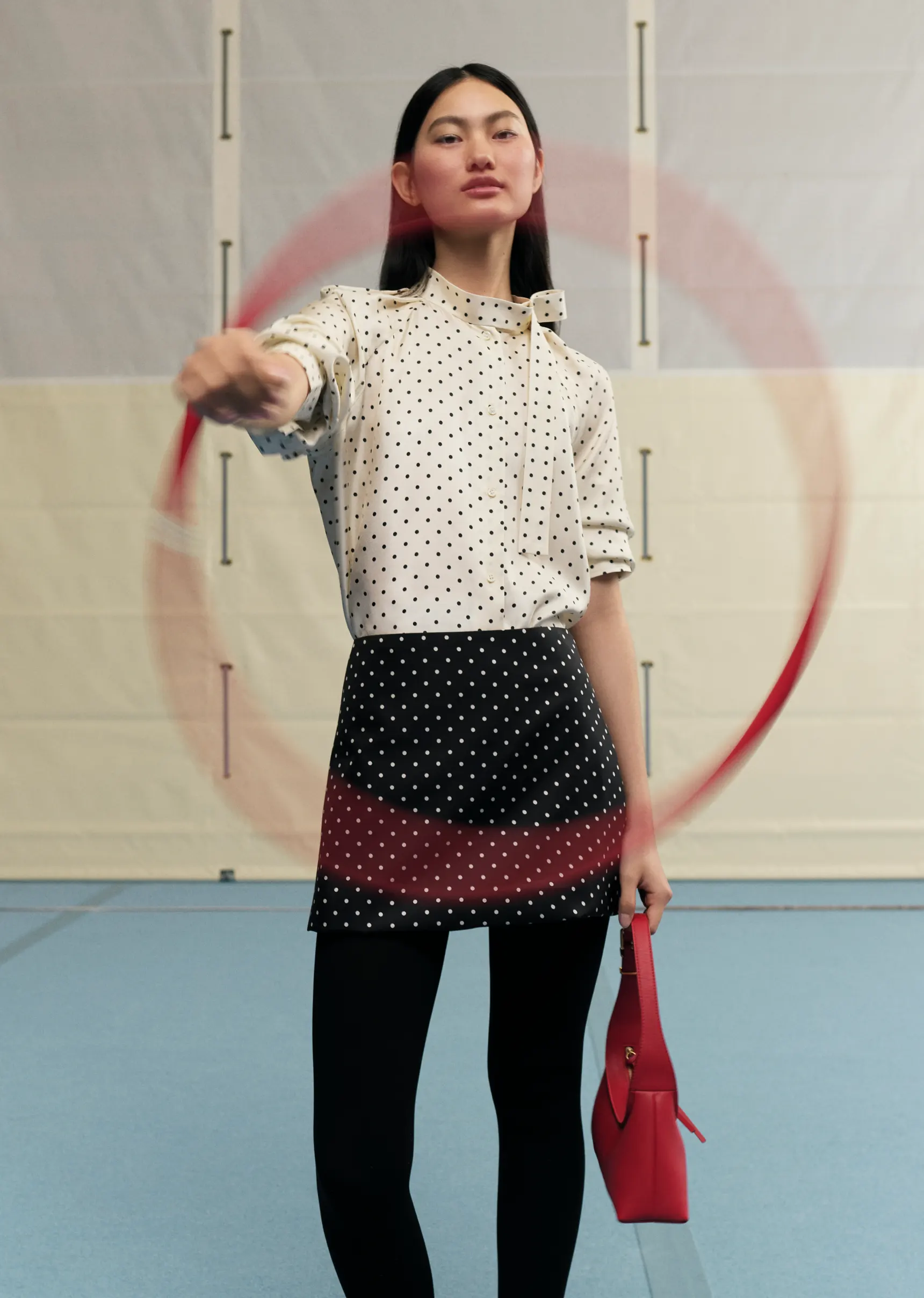 A fashionable young woman wearing a dotted top and skirt spins a red hula hoop in an indoor setting with a red purse.