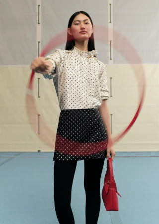 A fashionable young woman wearing a dotted top and skirt spins a red hula hoop in an indoor setting with a red purse.