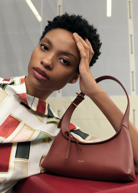 Brunette woman with a patterned shirt is seated indoors, resting her head on her hand next to a brown leather handbag on red cushion.