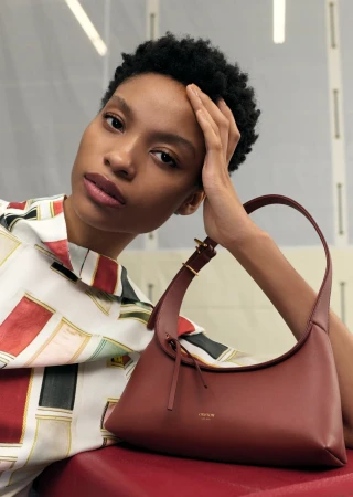Brunette woman with a patterned shirt is seated indoors, resting her head on her hand next to a brown leather handbag on red cushion.