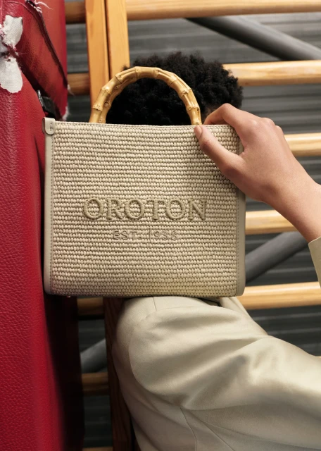 Close-up of a beige straw tote handbag with bamboo handles and the brand name 'OROTON' and 'EST. 1938' displayed, held by a person.