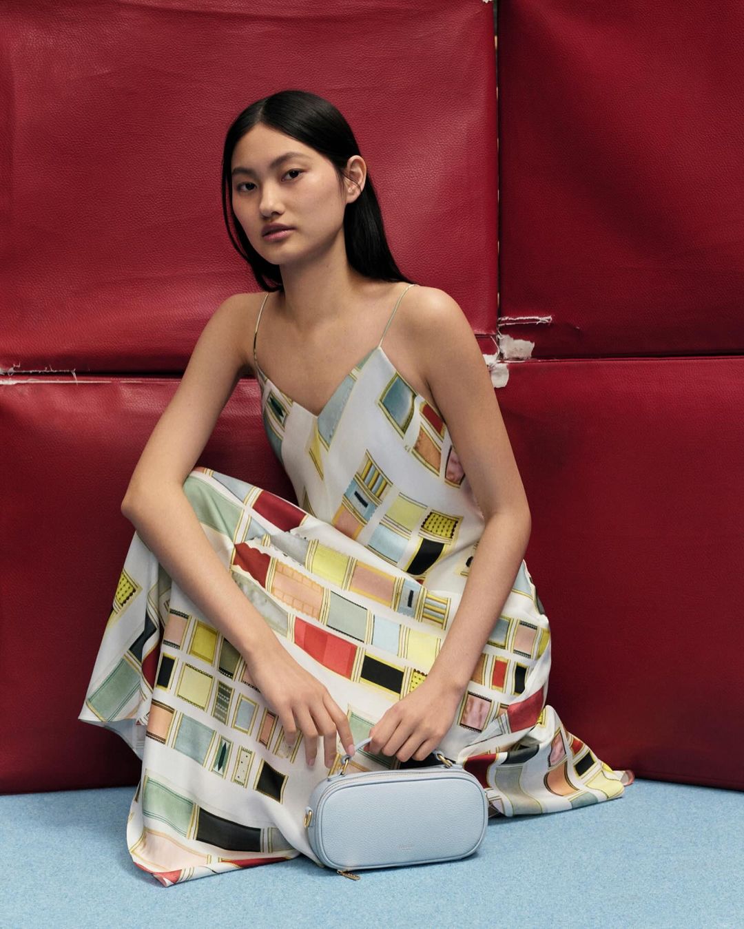 Asian woman in patterned slip dress sitting indoors with a light blue purse, posing with a neutral expression and soft studio lighting.