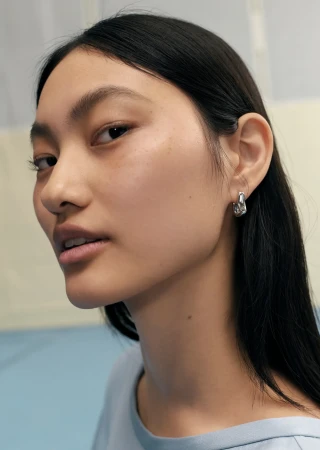 Close-up portrait of an East Asian woman wearing silver hoop earrings and a pale blue top against a minimal background.