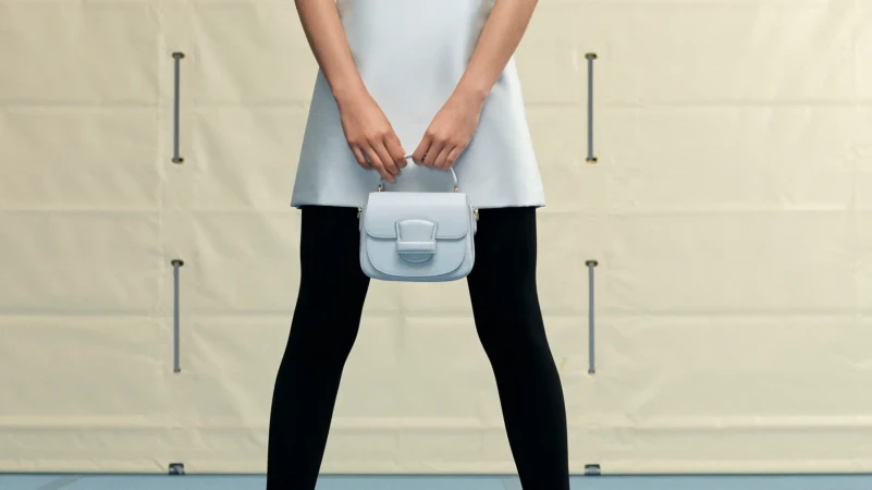 A light blue handbag is held in front of a fashion model wearing a white dress and black leggings against a neutral backdrop.