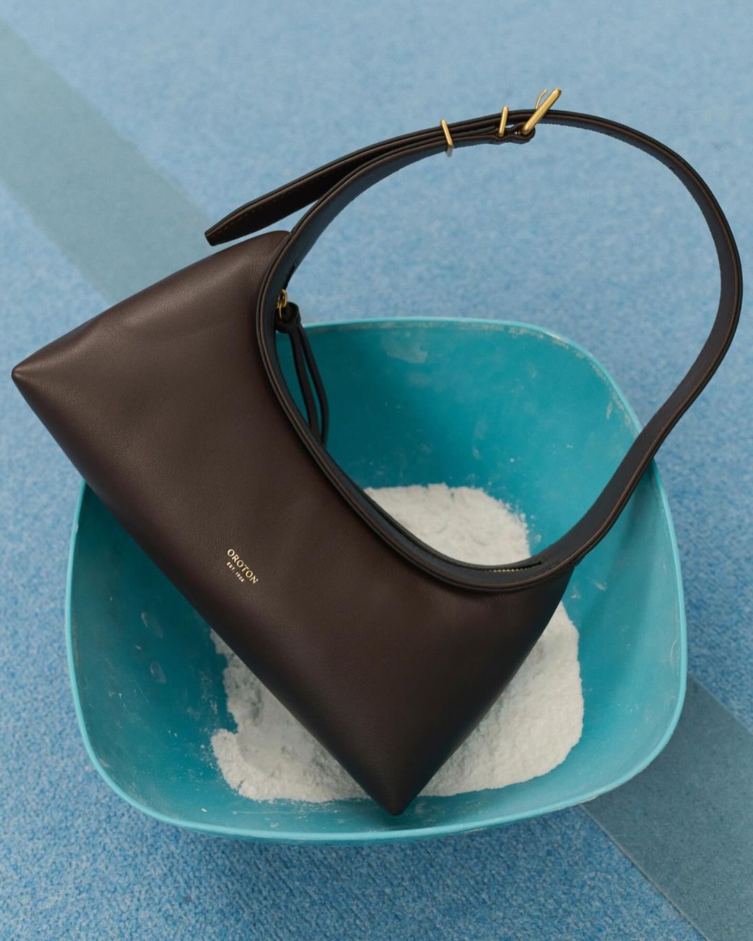 Brown leather shoulder bag on a blue surface with a turquoise bowl containing white powder. Studio product shot.