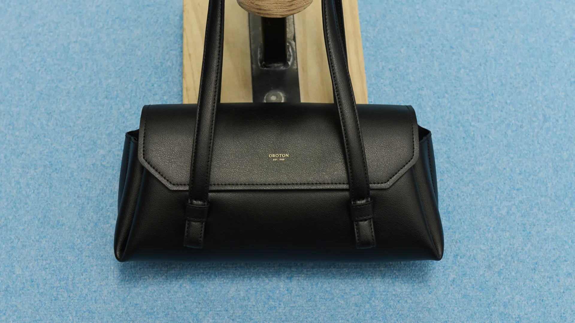 Black Oroton handbag with shoulder straps, centered against a blue textured background, showcasing fashion accessory details and studio lighting.