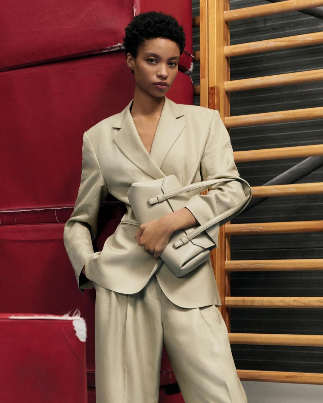 Fashion shot of an African-American woman in a light-beige suit holding a matching handbag, posing against a red and wood background.