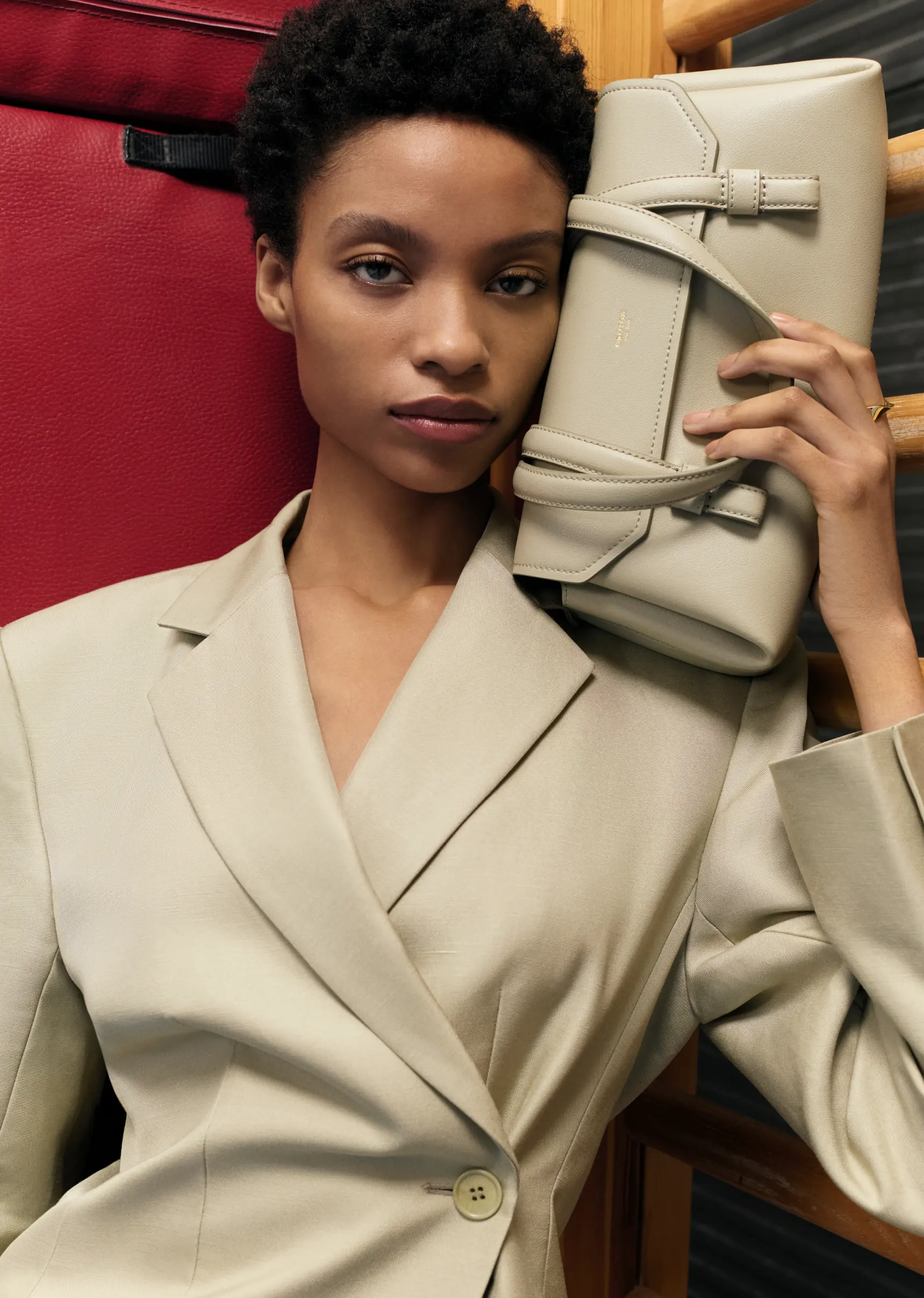 An adult female poses indoors with a light-beige satchel handbag, wearing a matching beige blazer with short natural dark hair.
